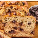A loaf of protein banana bread with chocolate chips on a wooden cutting board. A few slices have been cut off and are leaning against the loaf. Around the loaf is a bowl of chocolate chips, some chocolate chips and walnuts, a slice of banana bread, and some bananas.