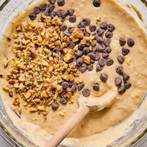 Bowl of banana bread batter with chocolate chips and walnuts sprinkled on top before they are mixed in. There is a wooden spoon in the bowl as well.