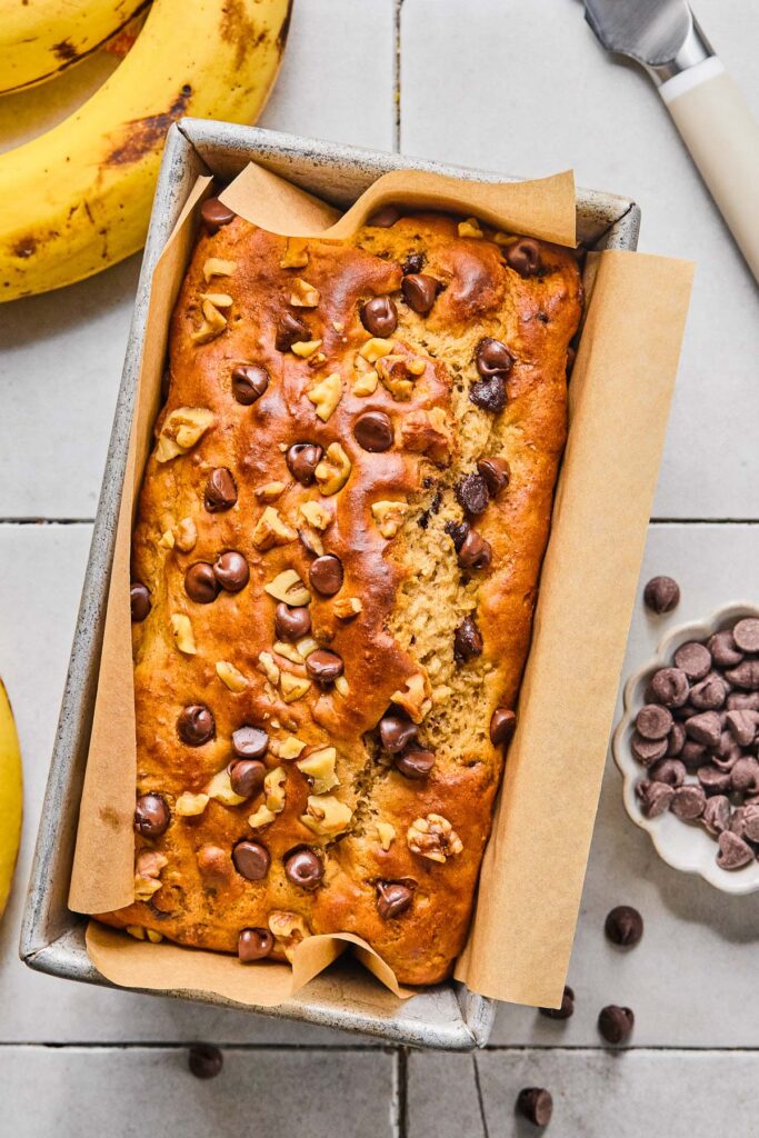 A loaf of high protein banana bread with chocolate chips and walnuts in a loaf pan. Around the pan is a bowl of chocolate chips, some bananas, and a knife.
