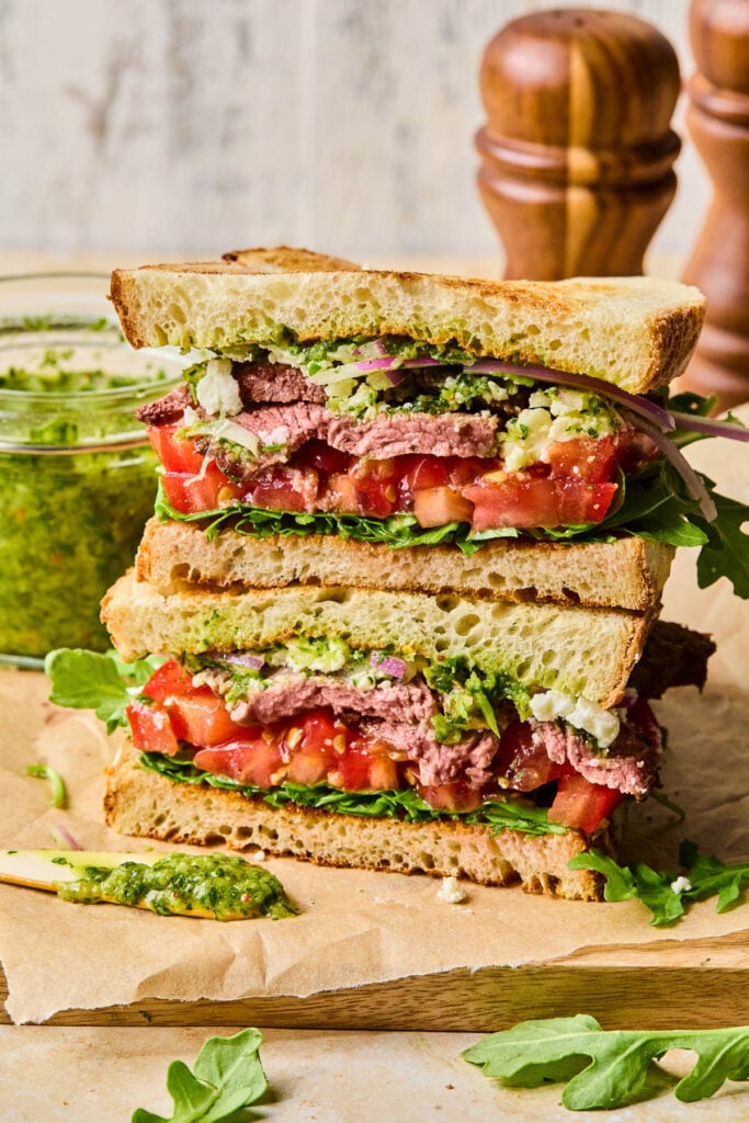 Two halves of a chimichurri steak sandwich with red onion, feta cheese, tomato, and arugula stacked on top of each other on a cutting board. Behind the sandwich is a jar of chimichurri and some wooden salt and pepper shakers.