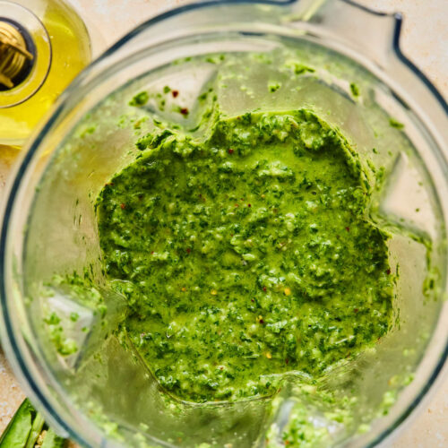 Blender chimichurri sauce in a blender. Around the blender is half a jalapeño, a jar of olive oil, and a juiced lime.
