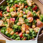 Kale caesar salad with chicken, veggies, and croutons in a bowl with a fork in it. Around the bowl is a small bowl of croutons, another bowl of salad, and a glass with dressing in it.