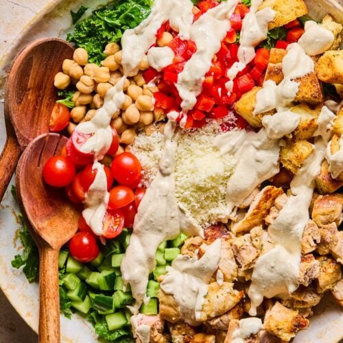 Large bowl with massaged kale, halved cherry tomatoes, chickpeas, diced red bell peppers, croutons, diced chicken thighs, diced cucumbers, and grated parmesan cheese all drizzled with dressing. Wooden salad servers are also in the bowl.