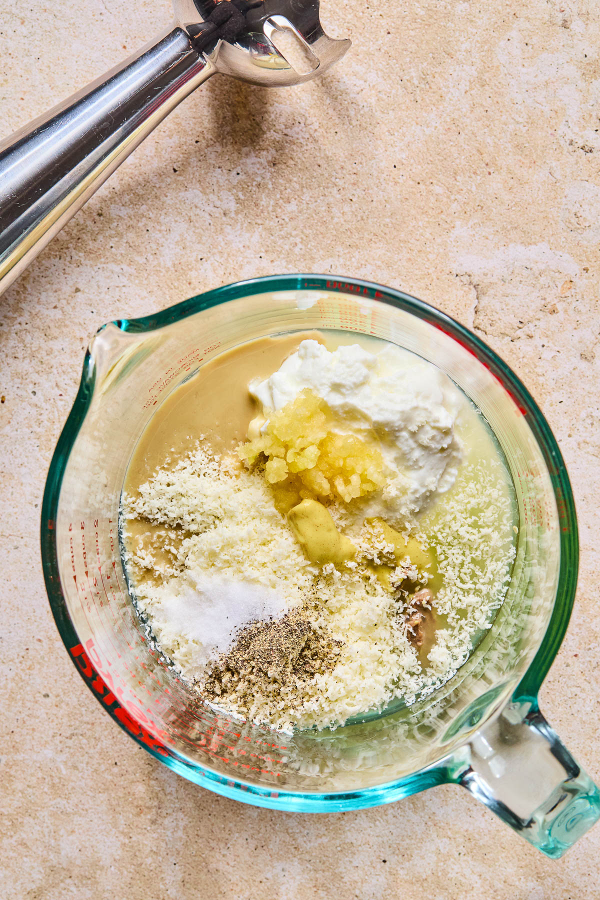Overhead shot of a measuring cup with greek yogurt, tahini, grated parmesan cheese, minced garlic, dijon mustard, anchovy paste, salt, and pepper in it. Next to it is an immersion blender.