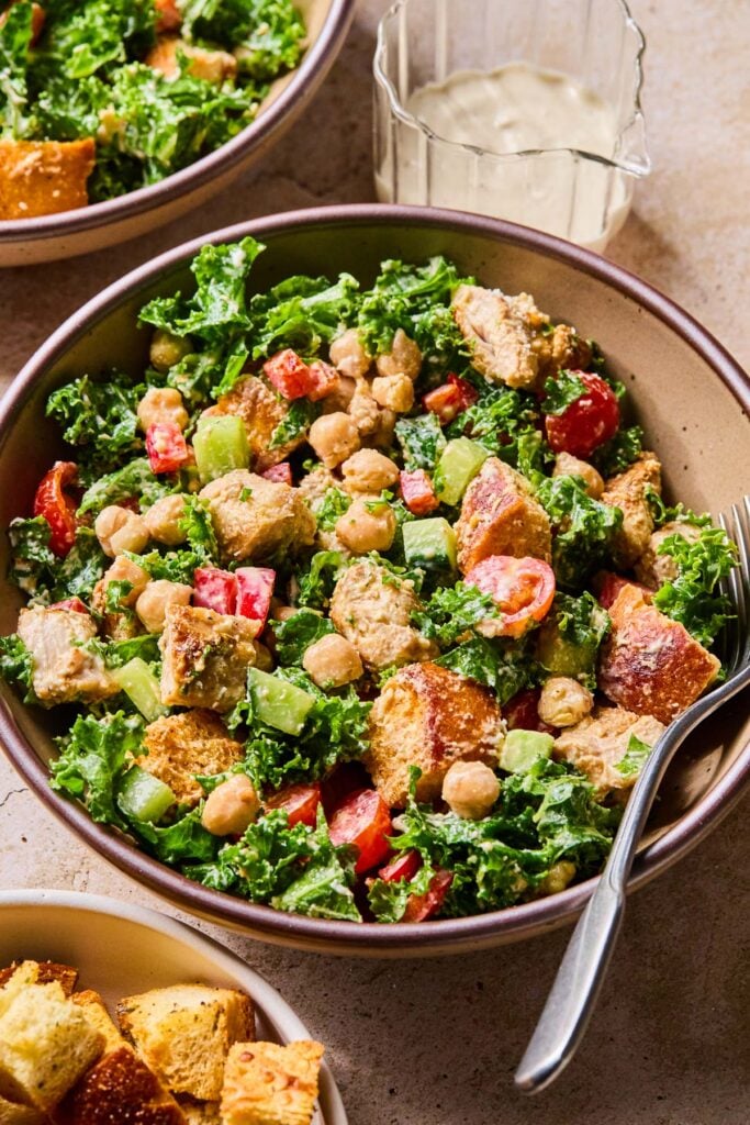 Kale caesar salad with chicken, veggies, and croutons in a bowl with a fork in it. Around the bowl is a small bowl of croutons, another bowl of salad, and a glass with dressing in it.