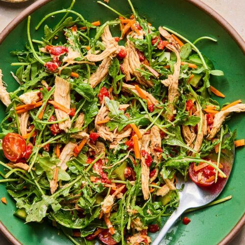 A shallow green bowl filled with a mix of arugula, shredded carrots, chopped cucumbers, halved cherry tomatoes, shredded chicken, and goat cheese tossed in olive oil and vinegar. There is also a fork in the bowl. Next to the bowl is a small bowl of salt and pepper.