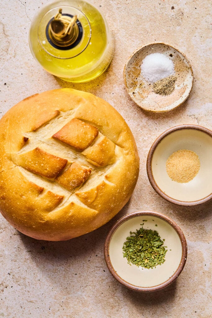 A small dish of salt, a small bowl of garlic powder, a small bowl of dried parsley, a load of bread, and a cruet of olive oil on a tan concrete counter.