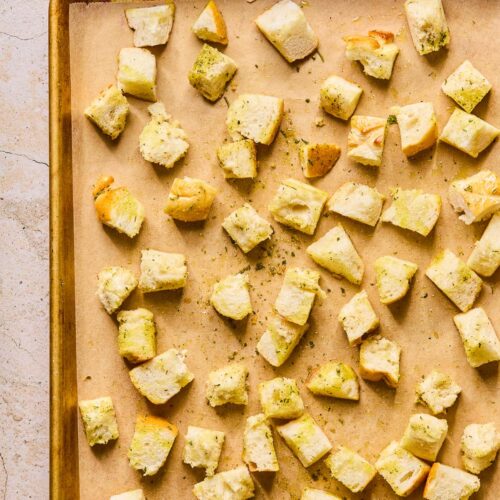 Cubed bread that has been tossed in oil and spices spread out on a parchment paper lined baking sheet. Next to the baking sheet is a cruet of olive oil.
