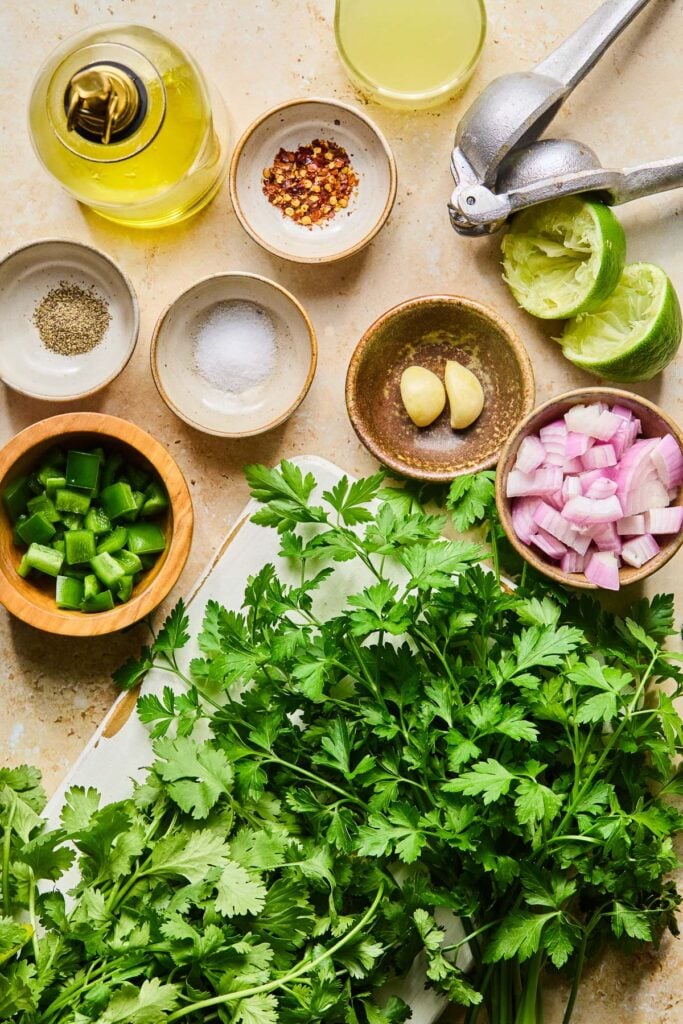 Small bowl of lime juice, a citrus squeezer, a lime that has been cut in half and juiced, a small bowl with two cloves of garlic in it, a small bowl of chopped shallot, a bunch of fresh parsley, a bunch of fresh cilantro, a small bowl of chopped jalapeño, a small bowl of salt, a small bowl of black pepper, a small bowl of red pepper flakes, and a jar of olive oil on a tan cement counter.
