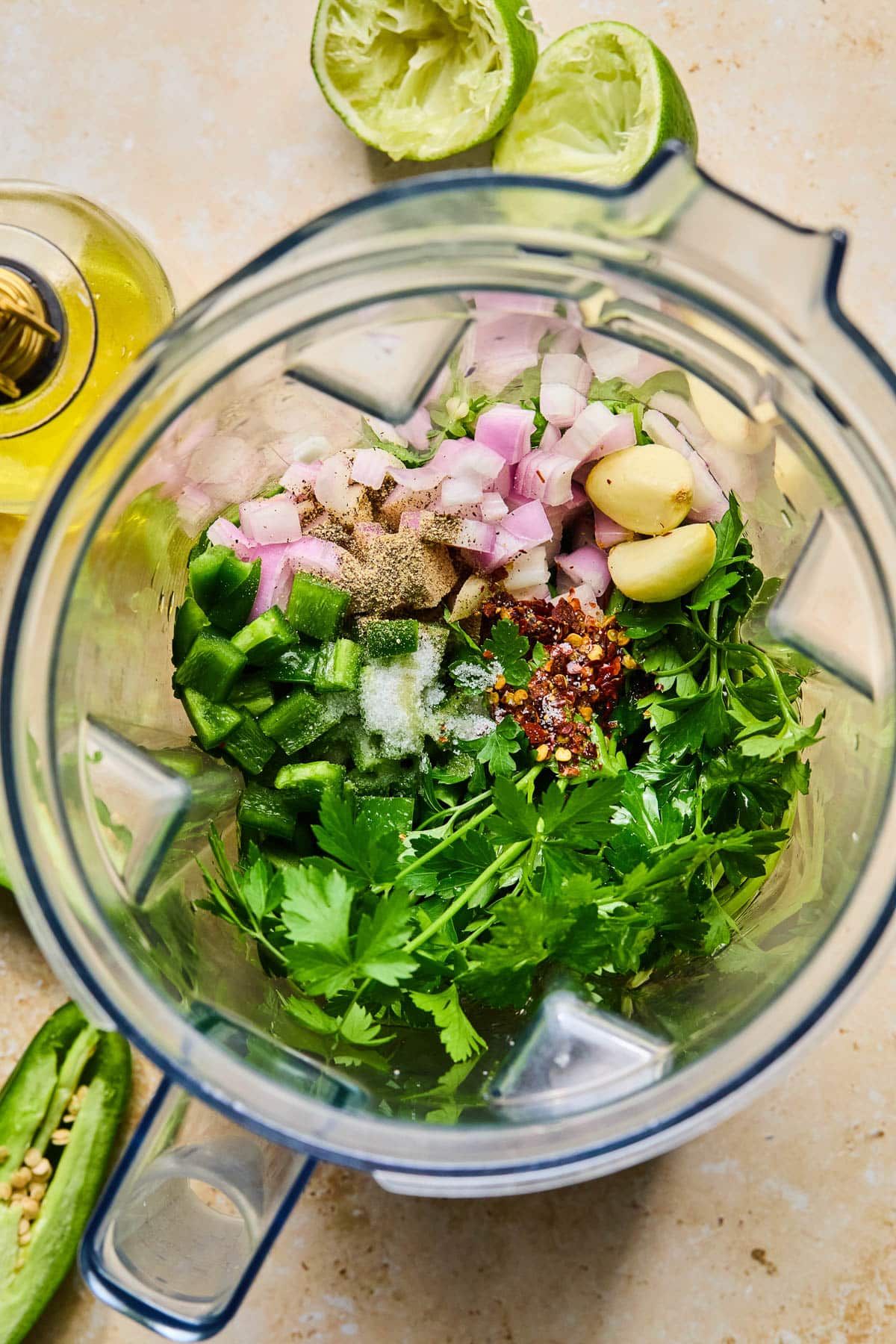 Blender will olive oil, fresh parsley, fresh cilantro, chopped jalapeño, chopped shallot, two garlic cloves, and spices before it is blended.