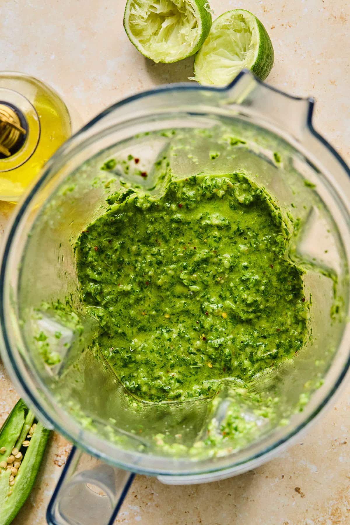 Blender chimichurri sauce in a blender. Around the blender is half a jalapeño, a jar of olive oil, and a juiced lime.
