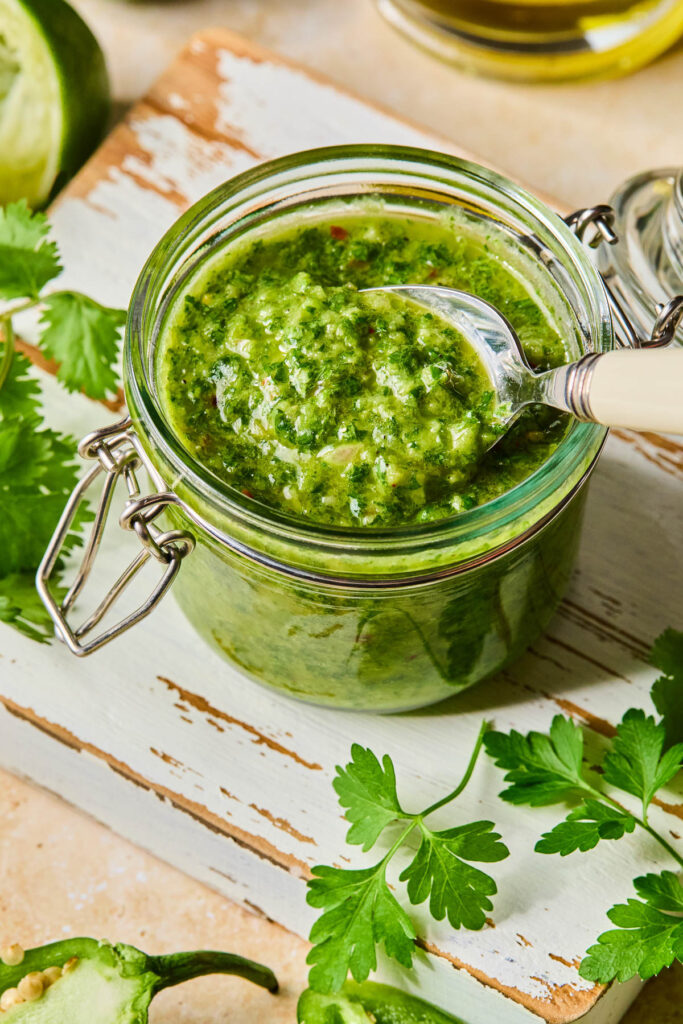 Jalapeño chimichurri in a jar with a spoon in it. The jar is on a white cutting board with fresh parsley leaves. Around the cutting board are a jalapeño, half a juiced lime, and a jar of olive oil.