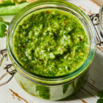A jar of spicy chimichurri sauce on a white wooden cutting board. Around the jar are some parsley leaves and a jalapeño that was cut in half lengthwise.