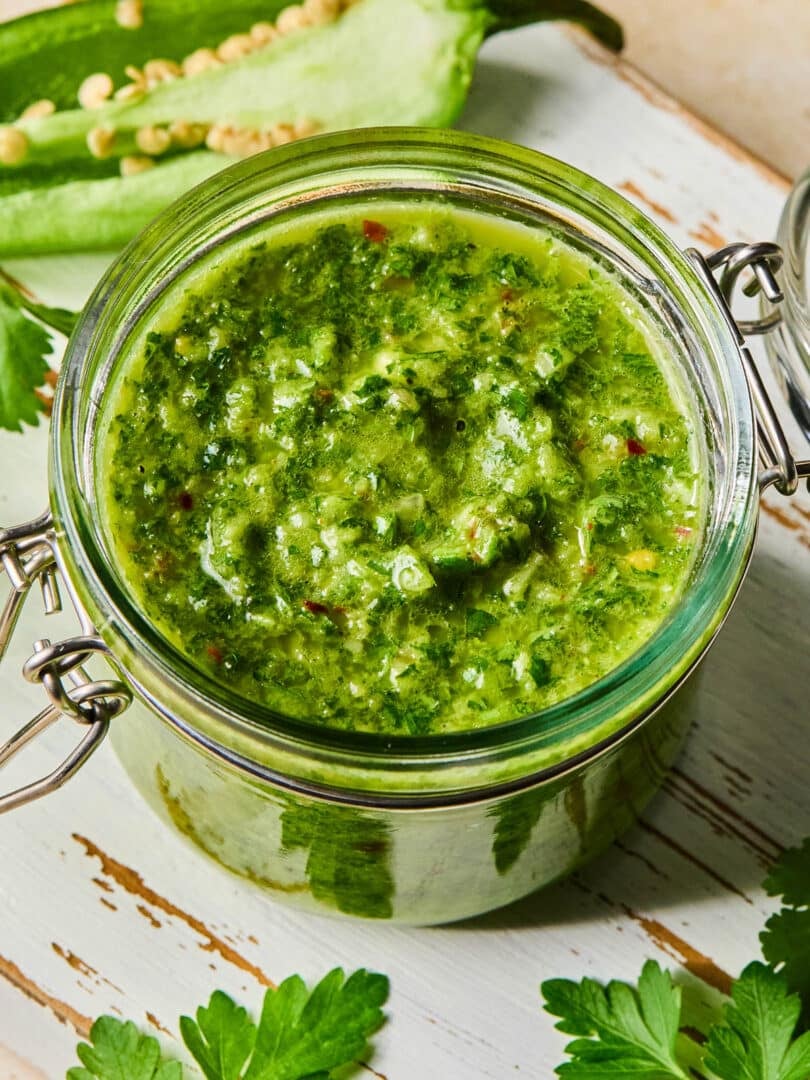 A jar of spicy chimichurri sauce on a white wooden cutting board. Around the jar are some parsley leaves and a jalapeño that was cut in half lengthwise.