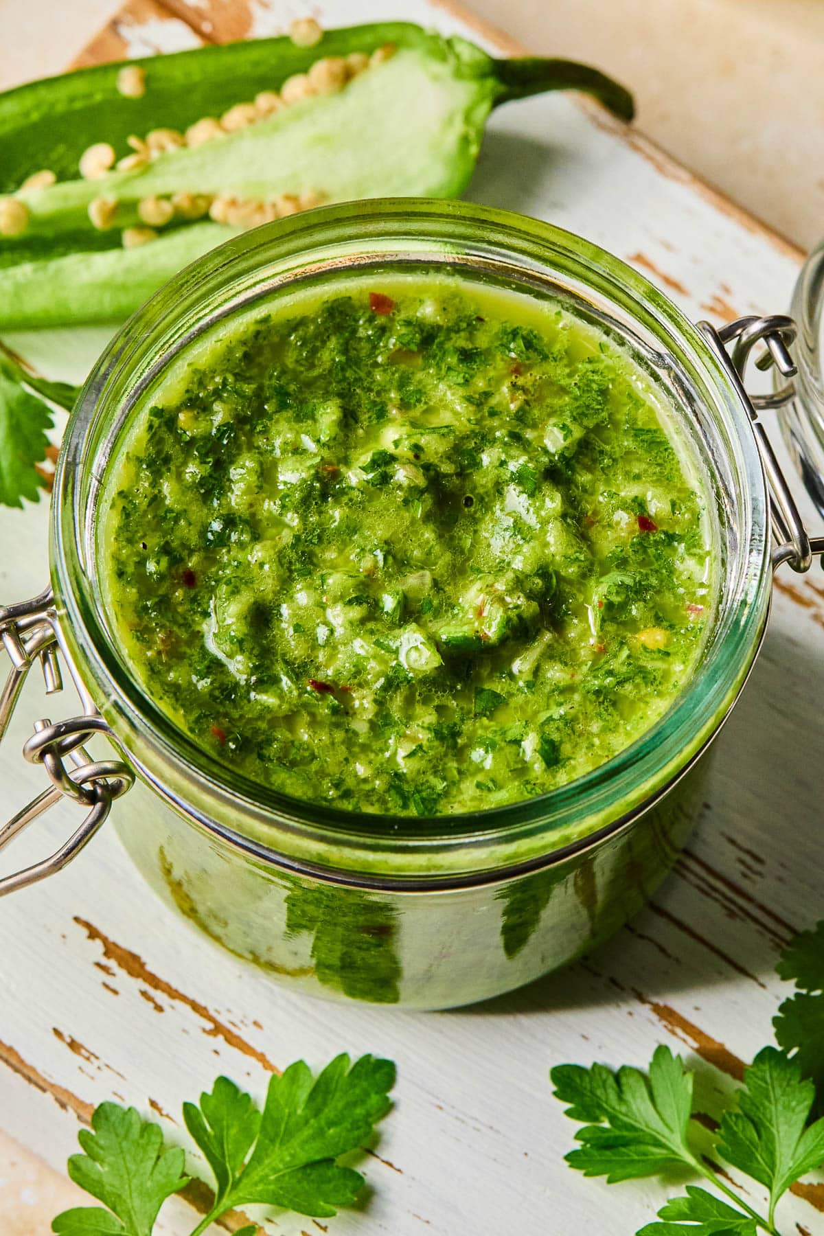 A jar of spicy chimichurri sauce on a white wooden cutting board. Around the jar are some parsley leaves and a jalapeño that was cut in half lengthwise.
