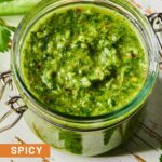A jar of spicy chimichurri sauce on a white wooden cutting board. Around the jar are some parsley leaves and a jalapeño that was cut in half lengthwise.